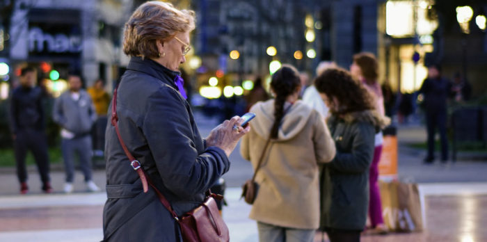 Ältere Frau steht an einem Fußgängerüberweg in der Nähe von Geschäften und tippt aktiv auf ihrem Smartphone herum.